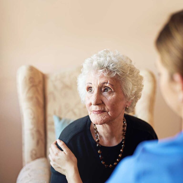 A senior resident receives her blood pressure checked by a caregiver in a bright, comfortable living space.